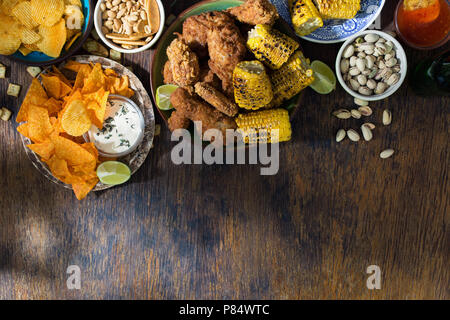 Fried chicken, sauces, potato chips, nachos, peanuts, pistachios and crackers on a wooden table with border, top view Stock Photo