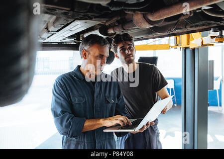 Auto technician using laptop while standing under the car with a mechanic. Two auto technicians working in auto service station. Stock Photo