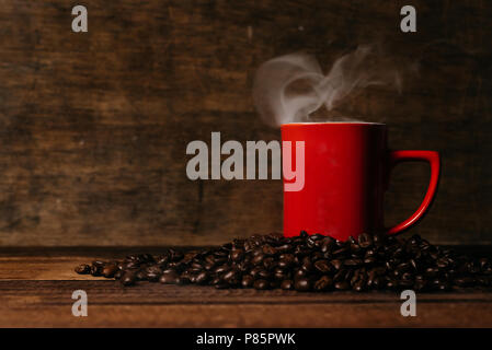 red mug with steam on a pile of fresh roasted coffee beans over wooden table. coffee break and beverage concept Stock Photo