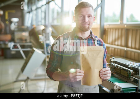 Waist up portrait of smiling bearded man wearing apron posing holding bag of coffee standing against roasting machines in artisan coffee roastery, cop Stock Photo