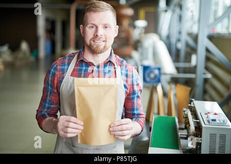 Waist up portrait of smiling bearded man wearing apron posing holding bag of coffee standing against roasting machines in craft  coffee roastery, copy Stock Photo