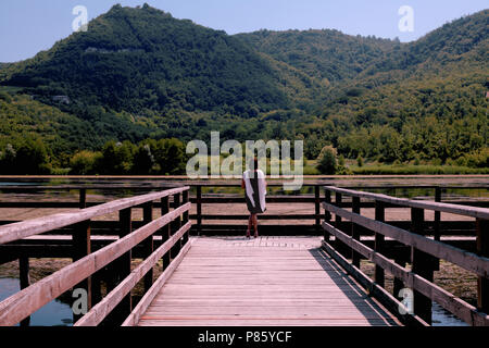 a woman with a white shawl is standing on the pier of the lake watching the green valley Stock Photo