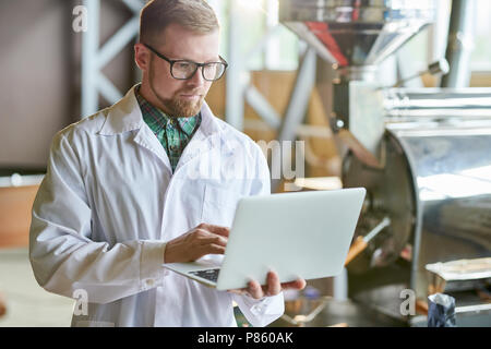 Waist up portrait of modern young man wearing lab coat using laptop while working in coffee production manufactory, copy space Stock Photo