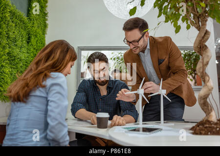 Smart qualified skilled young engineers looking at the project of alternative energy with miniatures of windmill turbines at the table. Stock Photo