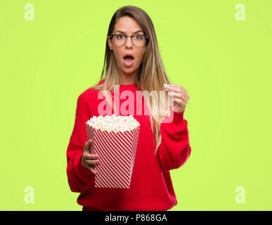 Beautiful young woman eating popcorn scared in shock with a surprise face, afraid and excited with fear expression Stock Photo