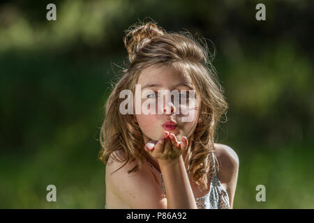 Posing in the sunshine, green background, young girl lblows kiss at camera, wearing a dress. Stock Photo