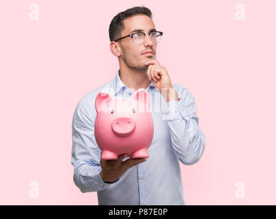 Handsome young man holding a piggy bank serious face thinking about question, very confused idea Stock Photo
