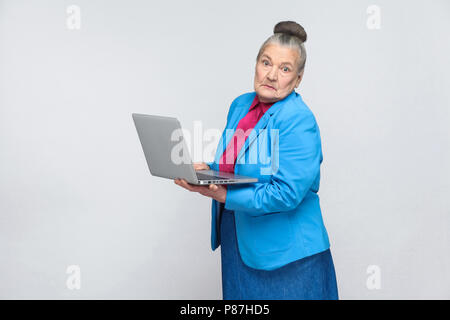 confused aged woman standing and holding laptop thinking and looking at camera. Grandmother in light blue suit with collected gray hair bun hairstyle. Stock Photo