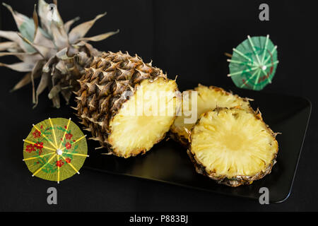 Sliced ripe and yellow pineapple in black plate and decorations for cocktails, umbrellas on dark background Stock Photo