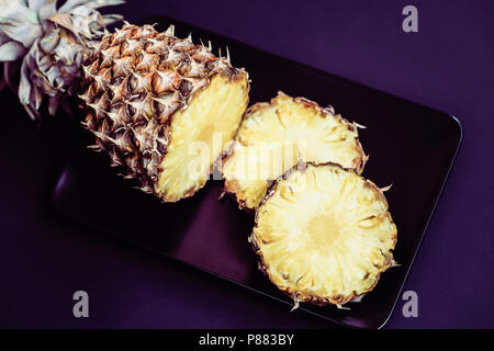 Slices of ripe pineapple on a black plate with decorations for cocktails, umbrellas. Dark violet background Stock Photo
