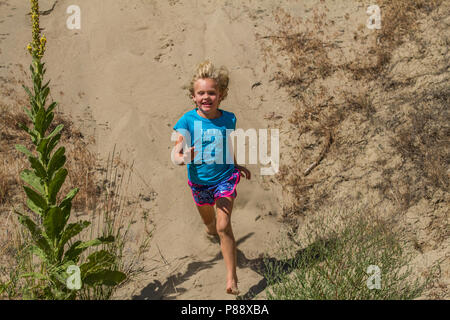 Young blonde girl, running in the sand,  laughing, hair and sand flying, having fun Stock Photo