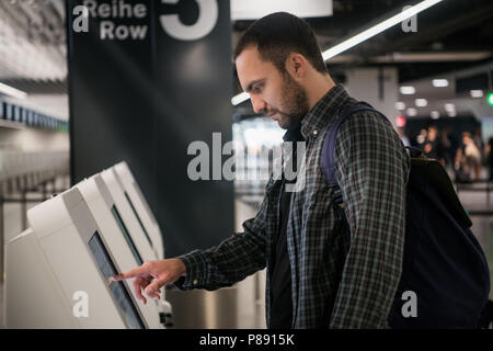 Young man with backpack touching interactive display using self service machine, doing self-check-in for flight or buying airplane tickets at automatic device in modern airport terminal building Stock Photo