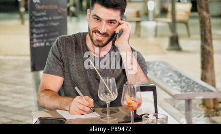 Young handsome man outside sitting at bar table in a summer day in city, calling someone on smartphone and writing with pen on paper sheet Stock Photo