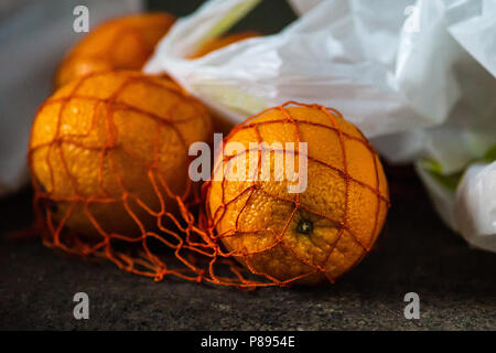 Fresh orange fruits in a string bag lay on a grunge stone surface. White plastic bag in the background. Stock Photo