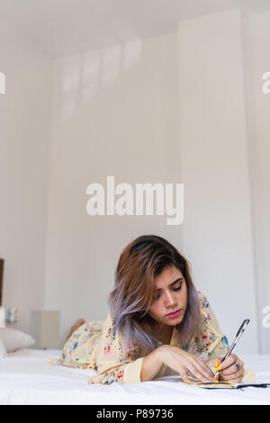 Lovely left-handed female making notes in small notebook while lying on soft bed in cozy room Stock Photo