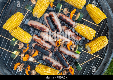 Sausages, corn on the cob and vegetable kebabs of mushroom, pepper and courgette cooking outside on a barbeque. Stock Photo