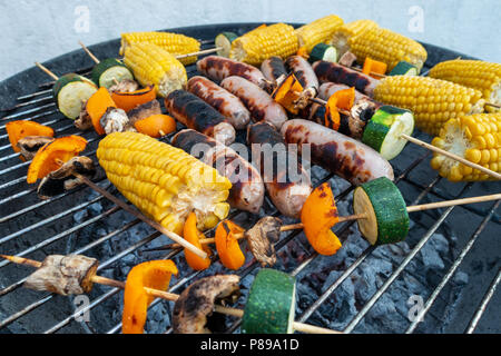 Sausages, corn on the cob and vegetable kebabs of mushroom, pepper and courgette cooking outside on a barbeque.