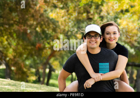 Young man carrying attractive young woman piggyback with colorful autumn leaves in distance Stock Photo