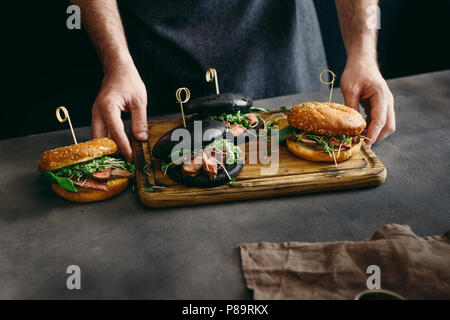 Man holding on wooden board different burgers with grilled beef meat. Dark style Stock Photo