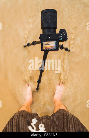 From above man standing with camera in wet coastal sand and shooting sunset in Bangkok, Thailand. Stock Photo