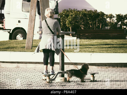 Elderly woman walking dog using outdoor exercise machine on city street in Spain Stock Photo