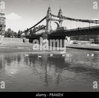 1950s, historical, Hammersmith Bridge, a view of the bridge on the Hammersmith riverside end. The bridge, which links the north side of the Thames to the south side, was the first suspension bridge over the River Thames when construction started in 1824. The bridge seen here is the second - replacement - one, built in 1887, which rests on the same pier foundations as the first. It was never designed to carry the weight and volume of road traffic which now exists in this part of inner London. Stock Photo