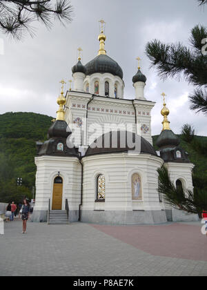 A beautiful church with white walls faces of saints with small domes with walking tourists standing on the background of a green slope behind it. . Fo Stock Photo