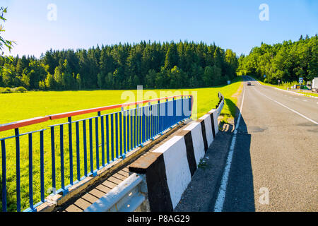 the end of the bridge, the asphalt road with the car passes along the sun-drenched grassy meadow through the dense green coniferous forest under the b Stock Photo