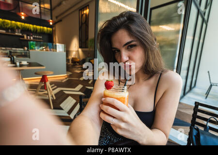 Attractive young woman holding glass of delicious smoothie and grimacing while sitting in nice cafe Stock Photo