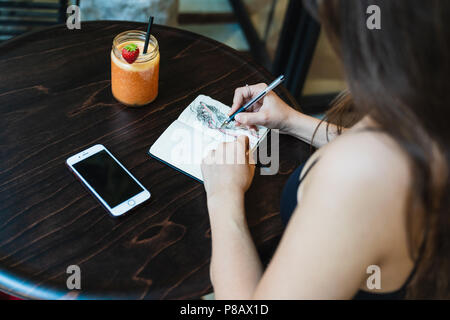 From above shot of anonymous young female sitting at cafe table and making sketches in small notebook Stock Photo