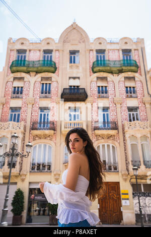 Charming young woman in casual outfit looking at camera while standing near building with beautifully decorated facade on street of Valencia in Spain Stock Photo