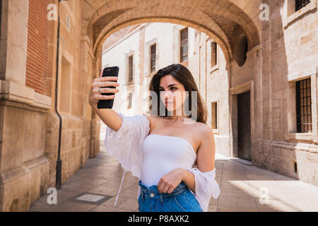 Beautiful young brunette holding smartphone and taking selfie while standing on street of Valencia on sunny day Stock Photo