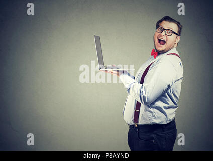 Side view of overweight business man in glasses holding laptop computer and looking shocked and amazed at camera on gray background Stock Photo