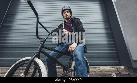low angle view of young man in helmet sitting on bmx bicycle and looking at camera on street Stock Photo
