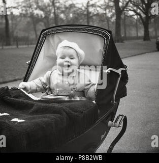 1950s, infant child in clothes and wearing a berry on their head sitting up in their coachbuit Royale luxury pram with a big chirpy smile on their face, England, UK. Stock Photo