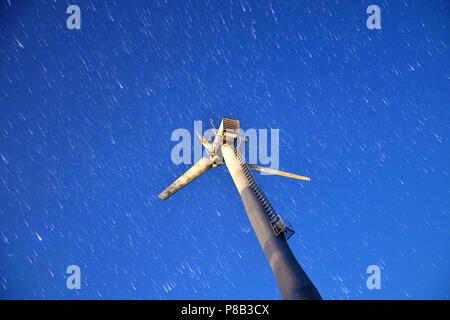 View from below of a damaged three blade wind turbine windmill over clear blue night sky with star trails. Stock Photo