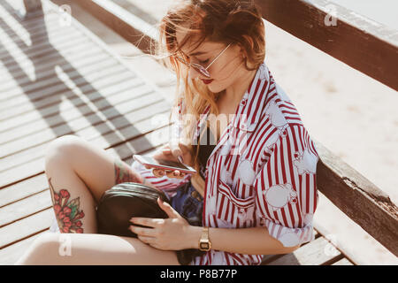 attractive young woman sitting on floor of wooden pier and using smartphone Stock Photo