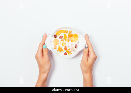 Woman's hands holding bowl of granola with milk, fresh fruits and nuts, top view. Healthy breakfast of oat muesli with yoghurt, hazelnuts and apricots Stock Photo