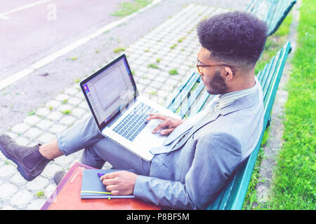 young contemporary black businessman sitting on a bench outdoor using laptop computer - portable office, remote working, business concept Stock Photo