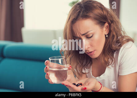 stressed depressed attractive young woman with painkiller pills and glass of water sitting on a sofa in her apartment Stock Photo