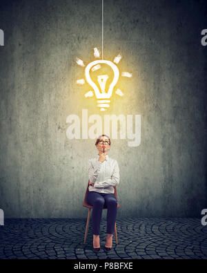 Stylish thinking businesswoman creating new strategy and looking up at bright light bulb while sitting on chair against gray background Stock Photo