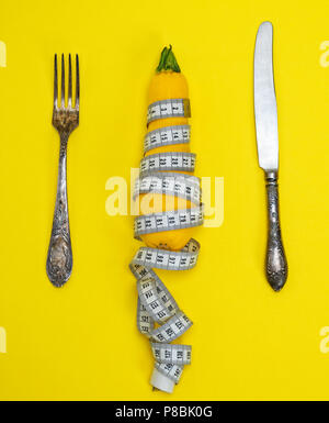 yellow zucchini wrapped in a measuring tape and an iron fork with a knife on a yellow background Stock Photo