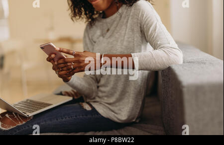 Businesswoman sitting on sofa at home using mobile phone while working on laptop. Woman working on laptop computer at home and managing her business. Stock Photo