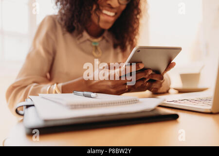 Smiling woman entrepreneur looking at a tablet pc with business documents and coffee on table while working on laptop. Businesswoman holding a tablet  Stock Photo