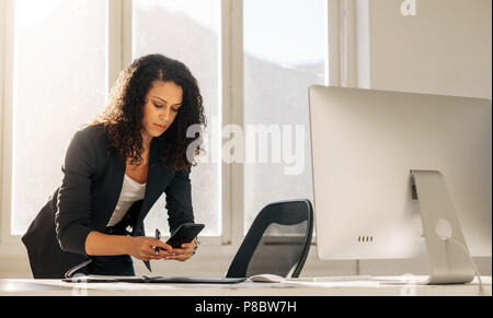 Businesswoman checking her mobile phone holding a pen to make notes leaning over her table. Woman entrepreneur using cell phone while making notes sta Stock Photo