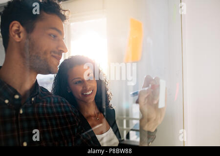 Office colleagues discussing business ideas and plans on a transparent glass board. Business couple writing on sticky notes pasted on glass board in o Stock Photo
