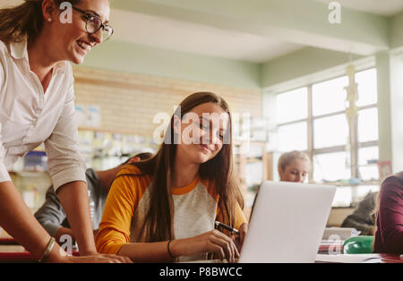 Young woman studying on laptop with teacher standing by in classroom. High school teacher helping student in classroom. Stock Photo