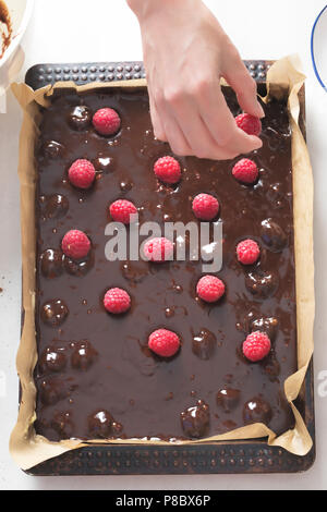 Woman placing raspberries on chocolate brownie batter cake in baking tray with parchment paper Stock Photo