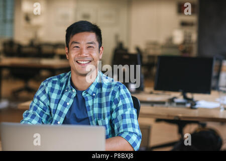 Smiling young Asian designer using a laptop at his desk Stock Photo
