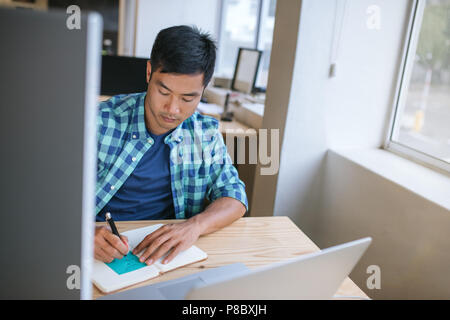 Young Asian designer sitting at his workstation writing down notes Stock Photo